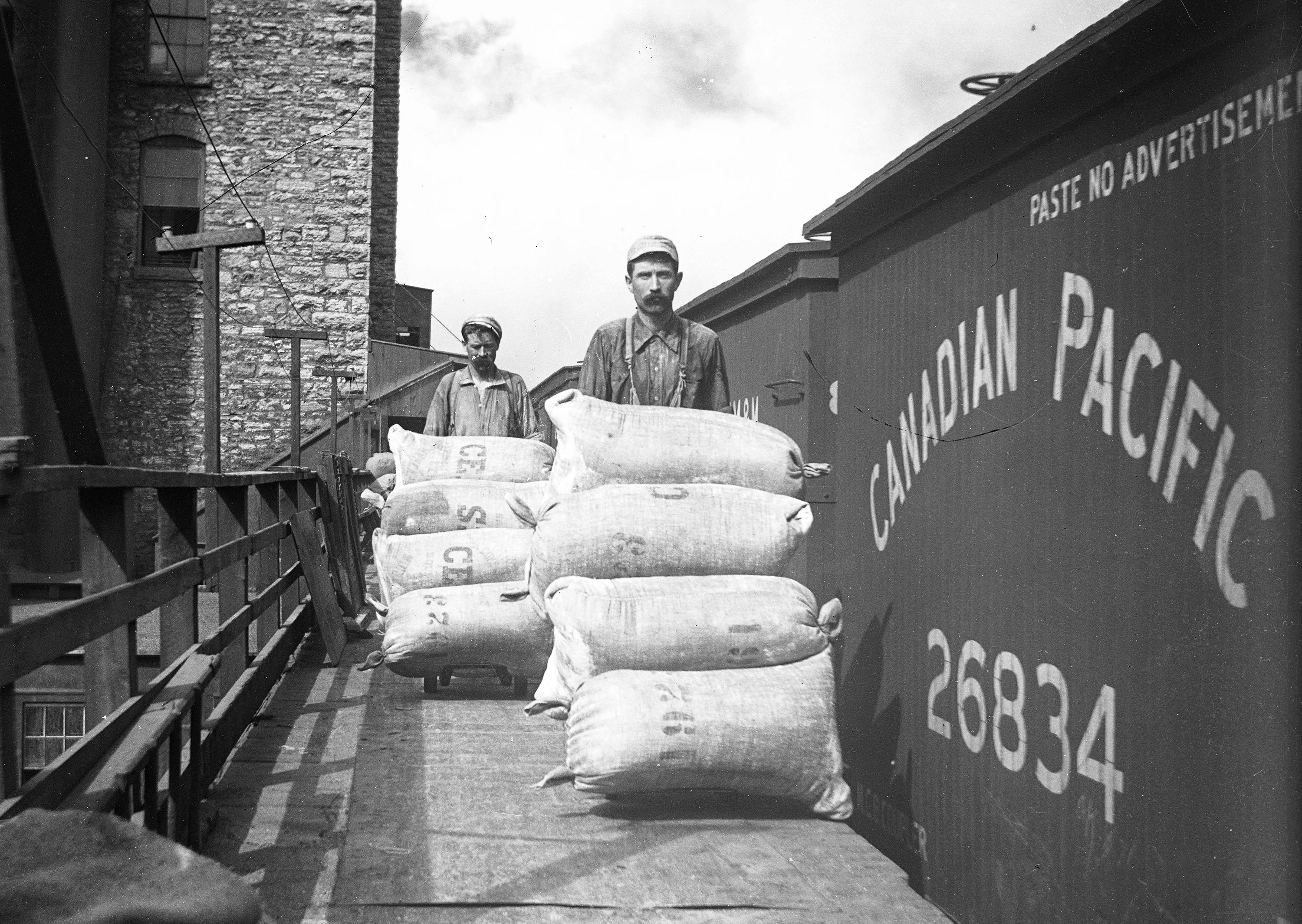 Workers load sacks of flour onto rail cars, likely around the turn of the century.