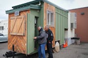 Michael Goze, left, and David Goodman peek around the outside of a tiny home trailer that was donated to their nonprofit which is sitting outside the 