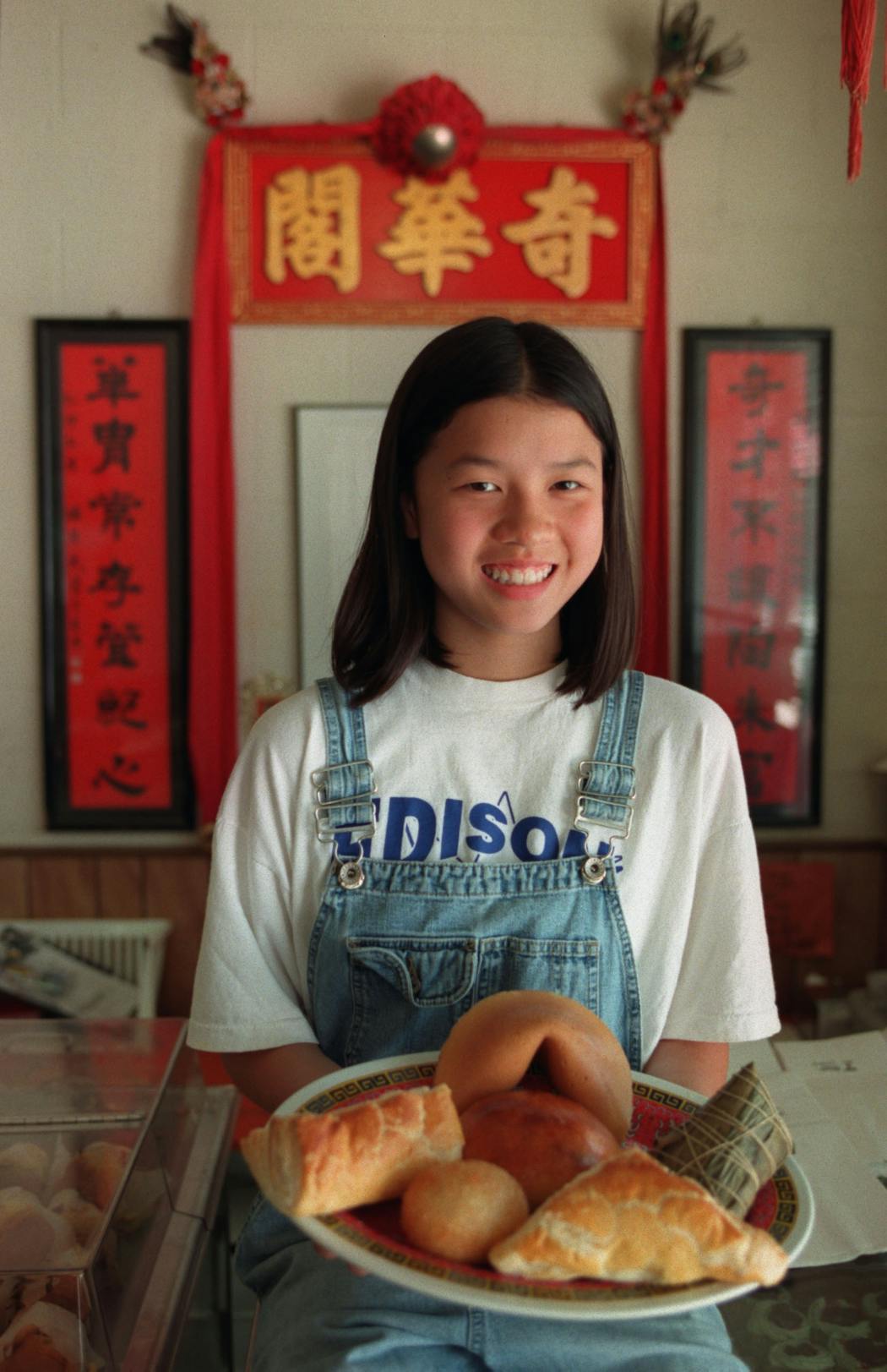 In 1998, Michelle Kwan held up a plate of baked goods from her family’s bakery. The items are, beginning with the giant fortune cookie at rear and moving clockwise: the big cookie, sticky rice wrapped in a banana leaf, curry puff pastry, sticky rice, barbequed pork puff pastry, and in the center, a curry bun.